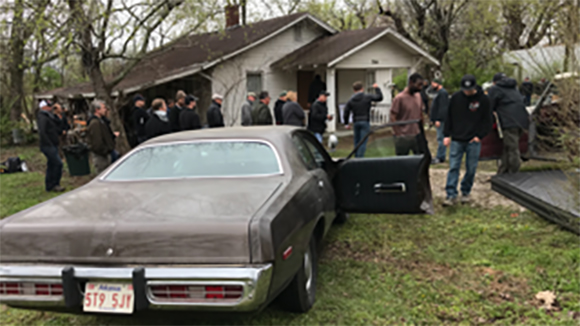 A group of people standing in a yard near a white house and a vintage brown coupe.