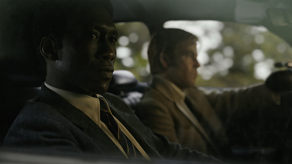 A Black man in a suit looks forward while a white man sits in the driver's seat of a car.