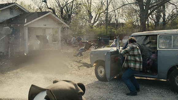 A person in a plaid shirt climbs into a light blue car while dust and debris fly in a yard.