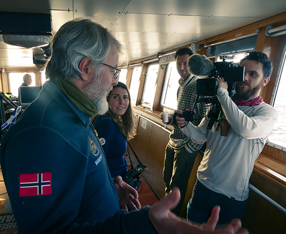 A man in a blue jacket talks to a woman and a cameraman filming him inside a boat cabin.