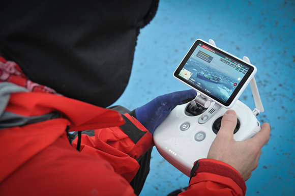 A person in a red jacket holds a white drone controller with a screen displaying a live video feed.