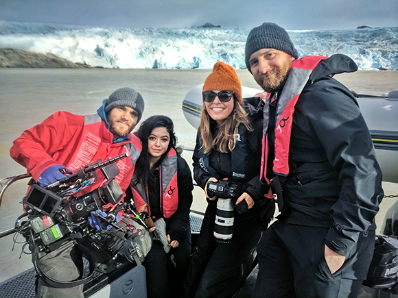 Four people in winter gear pose together with camera equipment in front of a large glacier.