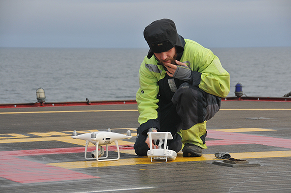 A person in a lime green jacket crouches on a ship deck, preparing a white quadcopter drone.