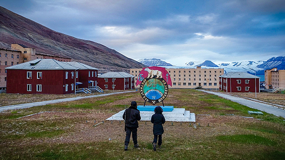 Two people stand before a large circular monument featuring a polar bear in a mountainous Arctic landscape.
