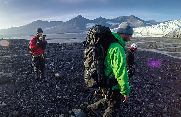 Hikers with large backpacks walk across a rocky terrain toward a glacier and mountains.