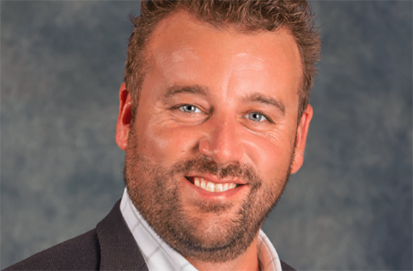 A smiling man with short brown hair, blue eyes, and a groomed beard wearing a dark blazer and white shirt.