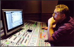 A man sits at a large audio mixing desk, looking intently at a computer monitor displaying recording software.