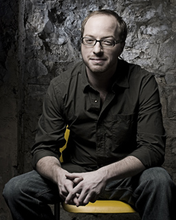 A man with glasses and a dark button-down shirt sits on a yellow chair against a stone wall.