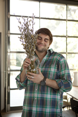 A man wearing a green and blue plaid shirt holds a glass vase filled with branches, smelling them with his eyes closed.