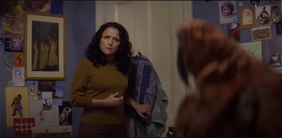 A woman with dark wavy hair wearing a mustard sweater looks toward a blurry figure in the foreground.