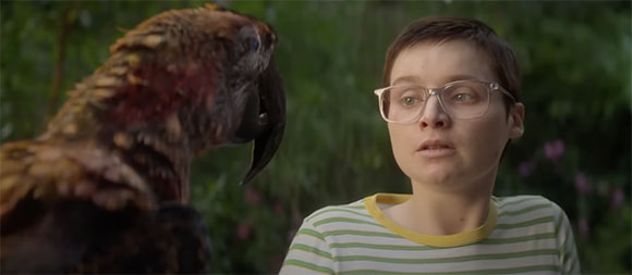 A person with short hair and glasses looks wide-eyed at a large dark parrot in a garden.