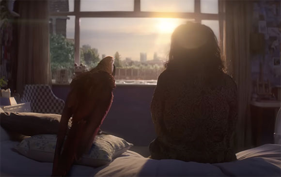A person and a large bird sit on a bed, looking out a window at a bright sunset over a city skyline.