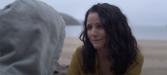 A woman with long dark wavy hair looks toward a person wearing a light-colored hood on a sandy beach.