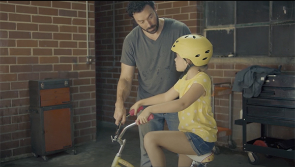 A man stands behind a young girl in a yellow shirt as she sits on a small bicycle in a workshop.