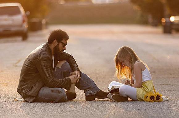 A man in a leather jacket and a young girl sit on a paved surface, facing one another.