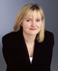 A blonde woman with blue eyes wearing a black blazer and a necklace against a gray background.