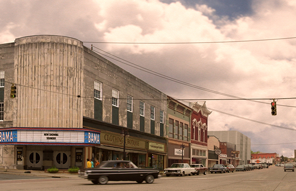 A street lined with multi-story commercial buildings and parked cars under a cloudy sky.