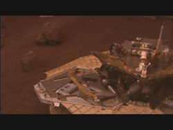 A robotic rover sits on a patterned landing platform amidst a dark, rocky, reddish landscape.