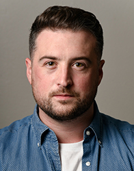 A man with short dark hair and a beard wearing a blue button-down shirt over a white t-shirt.