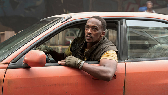 A man wearing a dark vest and gloves leans out of the driver's side window of an orange car.