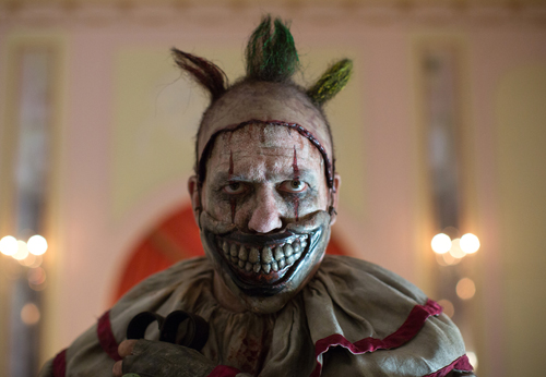 A person wearing disturbing clown makeup with large teeth and tufts of hair on their head.