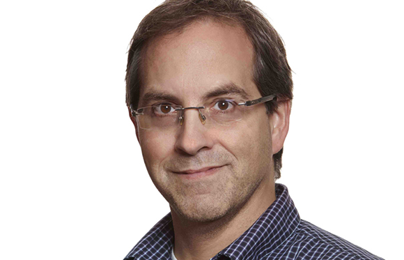 A man with short brown hair and glasses smiling slightly against a white background.