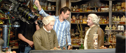 A man in a striped shirt stands between two actors in 18th-century period costumes on a film set.