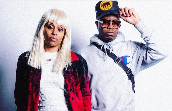 A woman with long blonde hair and a man wearing a black cap and sunglasses pose against a white background.