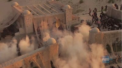 Dust clouds billow around stone buildings with domed towers in a desert setting.