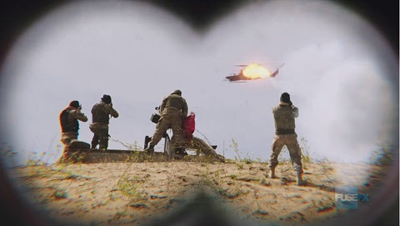 View through binoculars of four soldiers on a sandy hill watching a helicopter explode in the sky.