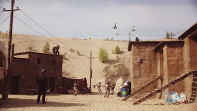 Military helicopters fly over a desert village with sand dunes and small buildings.