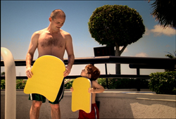 A shirtless man and a young boy stand side by side holding yellow swimming kickboards.