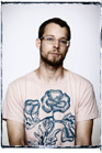 A man with short hair and a beard wearing glasses and a light pink t-shirt with a blue floral design.