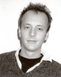 Black and white portrait of a man with short, spiked hair wearing a sweater over a collared shirt.