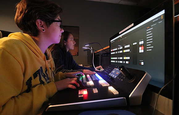 Two people sit at a desk using a professional video switcher and a large computer monitor.