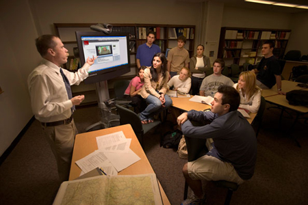 A man in a white shirt gestures toward a large monitor while students sit around tables listening.