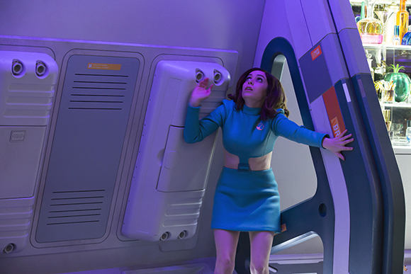 A woman in a teal two-piece outfit looks upward while standing in a futuristic, metallic corridor.