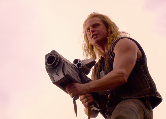 A man with long blonde hair holds a large, metallic sci-fi blaster against a pale pink sky.