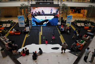 High-angle view of an indoor event area with a large screen displaying an orca and people sitting in lounge areas.