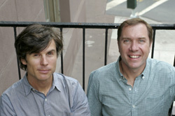 Two men in button-down shirts sit side-by-side outdoors near a black metal railing.