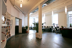 An open-plan office with white pillars, dark wood floors, bookshelves, and computer workstations.