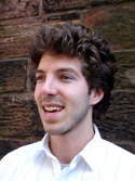 A man with curly brown hair and light stubble smiling in front of a stone wall.