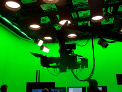 A large film camera on a crane arm positioned in front of a bright green screen studio backdrop.