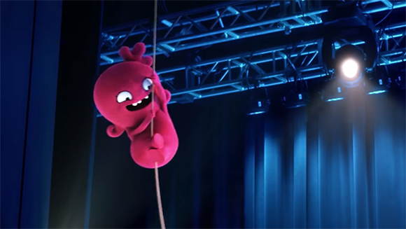 A bright pink, cartoonish creature hangs from a thin rope against a dark blue stage background.
