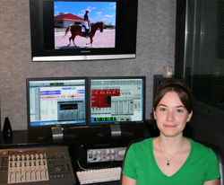 A woman in a green shirt sits before two computer monitors and a mixing console in a studio.