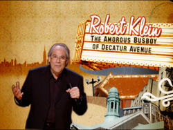 A man in a black suit gesturing while standing before a sign for 'Robert Klein: The Amorous Busboy of Decatur Avenue'.
