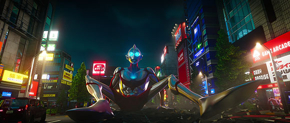 A giant silver humanoid figure sits in a brightly lit city street at night surrounded by tall buildings.