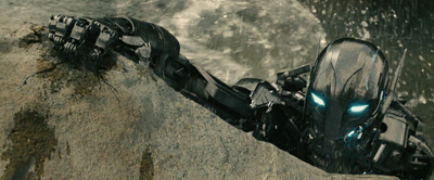 A dark, metallic robotic figure with glowing blue eyes climbs up a textured rock face during a rainstorm.