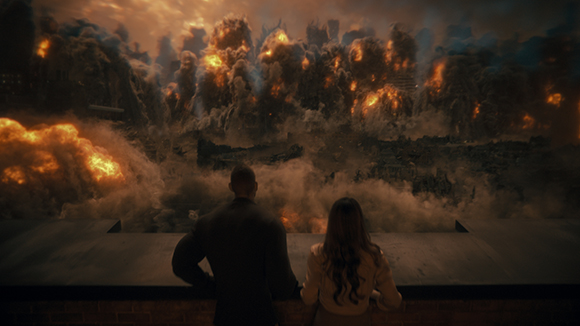 A man and woman from behind look out over a city engulfed in massive fireballs and thick smoke.