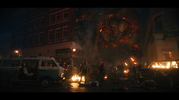 A massive, glowing fiery creature towers over a dark city street filled with smoke, fire, and people.